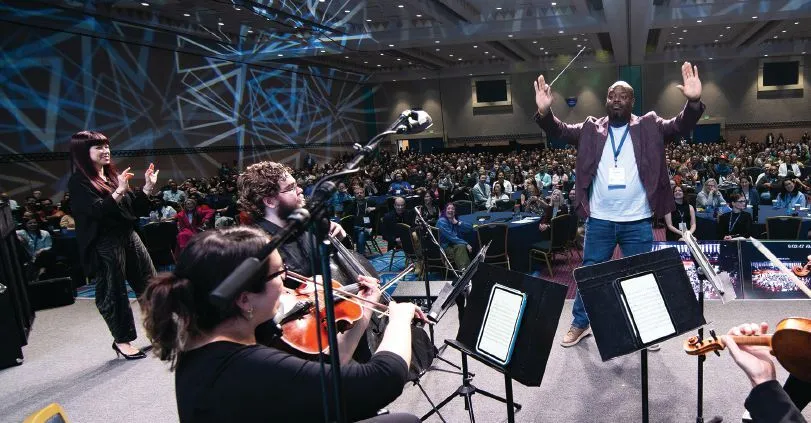 Attendee Artrell Williams tries his hand at conducting after a little coachingfrom keynoter Sarah Hicks (left). PHOTOGRAPHY BY HUGHES FIORETTI PHOTOGRAPHY